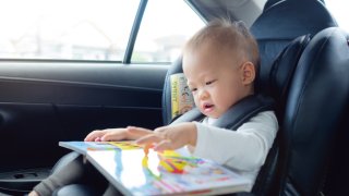 Cute little Asian 18 months / 1 year old toddler baby boy child sitting in car seat holding and enjoy reading book, Happy traveling with child concept, Little Traveler safety, road trip concept