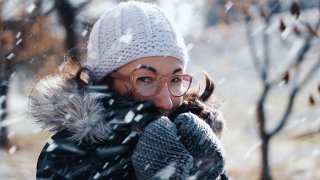 A woman wearing glasses and a beanie holds her gloved hands near her face and jacket as snow swirls around her.