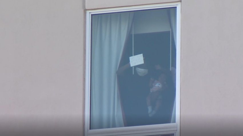 Una persona sujetando a un bebe frente a una ventana de hotel sujetando un papel que lee ayudame.